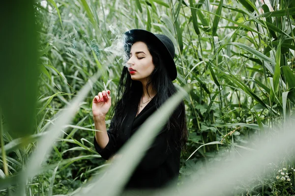 Sensual smoker girl all in black, red lips and hat. Goth dramatic woman smoking on common reed.