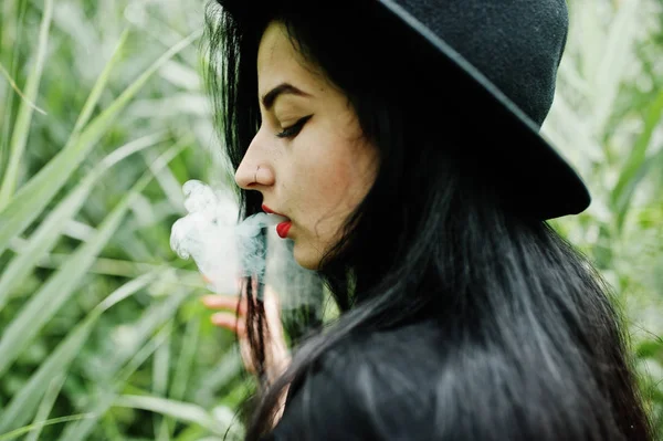 Sensual smoker girl all in black, red lips and hat. Goth dramatic woman smoking on common reed.