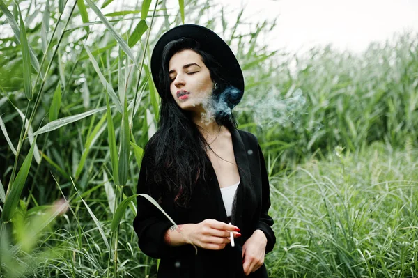 Sensual smoker girl all in black, red lips and hat. Goth dramatic woman smoking on common reed.