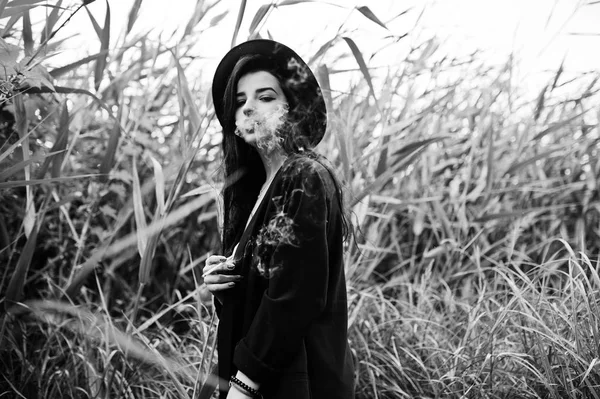 Sensual smoker girl all in black, red lips and hat. Goth dramatic woman smoking on common reed. Black and white portrait.