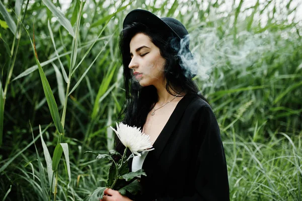 Sensual smoker girl all in black, red lips and hat. Goth dramatic woman hold white chrysanthemum flower and smoking on common reed.