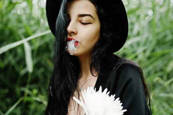 Sensual smoker girl all in black, red lips and hat. Goth dramatic woman hold white chrysanthemum flower and smoking on common reed.