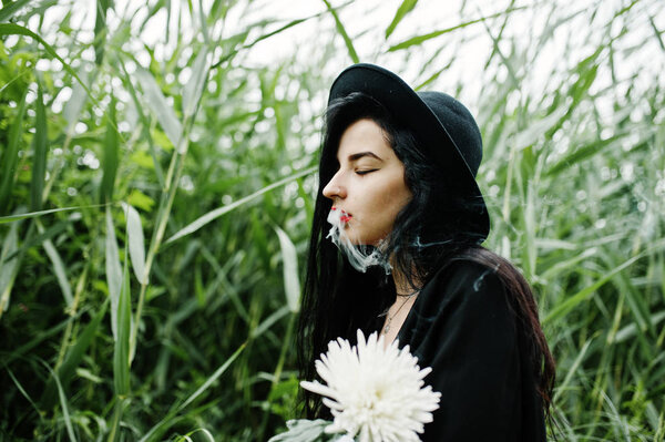 Sensual smoker girl all in black, red lips and hat. Goth dramatic woman hold white chrysanthemum flower and smoking on common reed.