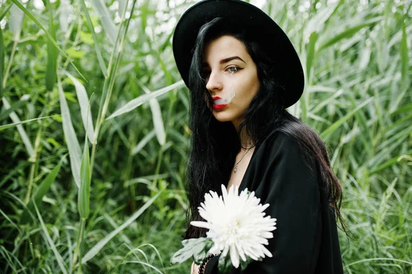 Sensual smoker girl all in black, red lips and hat. Goth dramatic woman hold white chrysanthemum flower and smoking on common reed.