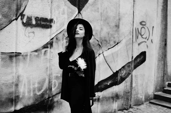 すべて黒で官能的な女の子は 赤唇し 帽子します ゴス劇的な女性は 落書きの壁に白い菊の花を保持します 黒と白の肖像画 — ストック写真
