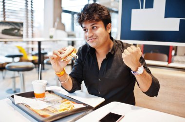 Fast food café'de oturan ve hamburger, yemek şık Hintli adam cep telefonu sabah haberleri okuyun. O mutlu ve onun gibi bir şey kazanmak göster.