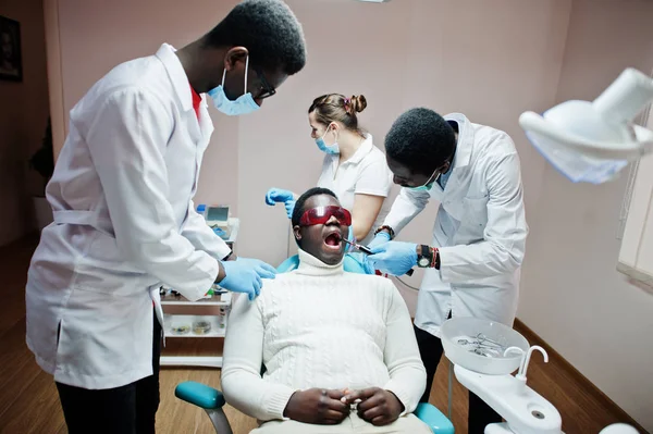 Multiracial dentist doctors team. African american man patient at UV protective glasses. His teeth treated with the help of a dental UV curing light lamp and a dental mirror.