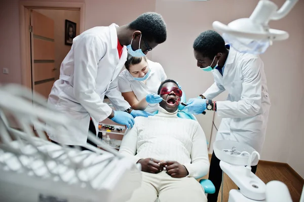 Multiracial dentist doctors team. African american man patient at UV protective glasses. His teeth treated with the help of a dental UV curing light lamp and a dental mirror.