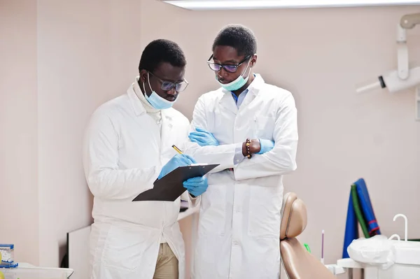 Two african american male colleagues doctors working together, discussing and looking at clipboard on clinic.