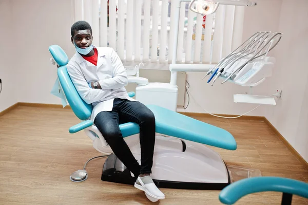 African american male doctor in mask with crossed arms sitting at dentist chair in dental clinic.