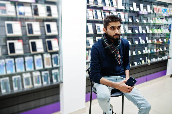 Indian beard man customer buyer at mobile phone store sitting on chair with smartphone at hand. South asian peoples and technologies concept. Cellphone shop.