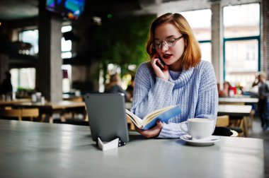Neşeli genç güzel redhaired kadın gözlük Cafe kahve ile üzerinde çalışma evinde otururken ona telefon, dokunmatik yüzey ve dizüstü bilgisayar kullanarak.