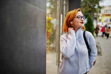 Neşeli genç güzel redhaired kadın gözlük, sıcak mavi yün kazak sırt çantası ile açık poz verdi.