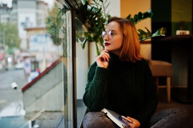 Güzel bir şey hayal. Neşeli genç güzel redhaired kadında defterini, pencere karşı kafe otururken kullanarak gözlük, yeşil sıcak yün kazak,.