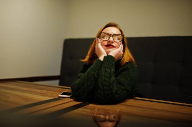 Neşeli genç güzel redhaired kadın gözlük, giymek yeşil sıcak yün kazak, kafede oturan üzerinde.