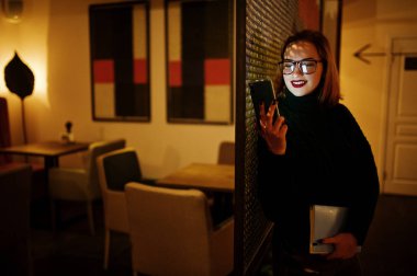 Cep telefonunuzun karanlık oda bardaklarda neşeli genç güzel redhaired kadın. Diisplaying belgili tanımlık telefon perde yüzünde.