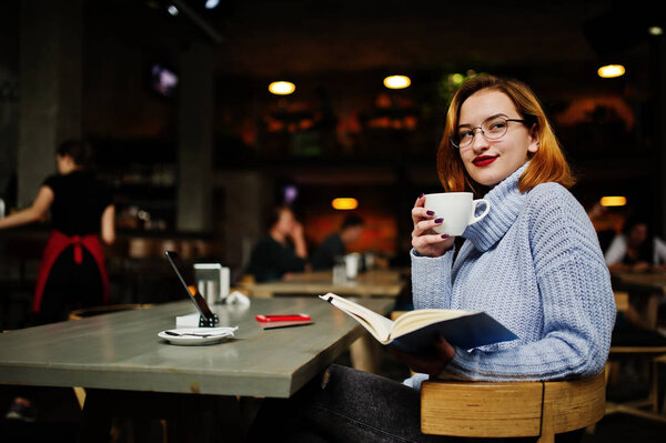 Веселая молодая красивая рыжая женщина в очках, используя свой телефон, тачпад и ноутбук, сидя на своем рабочем месте в кафе с чашкой кофе
.