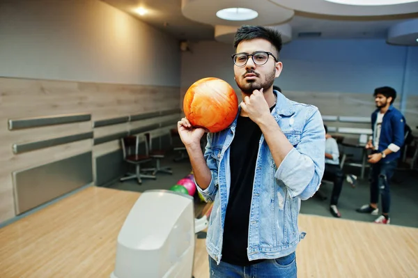 Stylish asian man in jeans jacket and glasses standing at bowling alley with ball at hand.