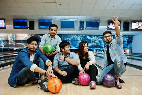 Group of five south asian peoples having rest and fun at bowling club, sitting on bowling alley with balls on hands.