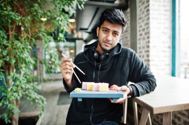 Rahat ve şık genç Asyalı adam suşi yemek kafede kulaklık ile.