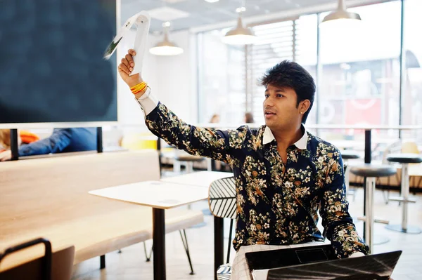 Young male indian freelancer sitting open laptop computer in fast food cafe, handsome asian man holds the plate with the order and waits for the waiter in comfortable coffee shop.