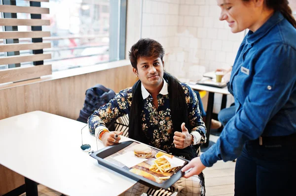 Girl waiter submits the tray with order burger and french fries to attractive and handsome stylish indian man wear on shirt in modern lounge cafe at his work break.
