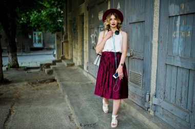 Şık ve güzel sarışın model kız şık Kırmızı kadife kadife etek, beyaz bluz ve şapka, telefon ve kulaklık ile poz verdi.