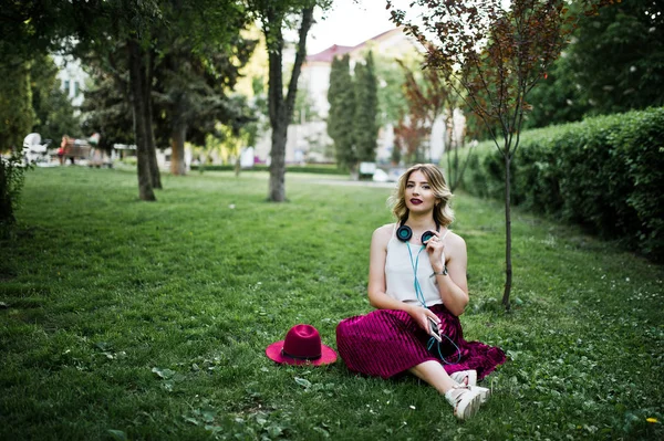 スタイリッシュな赤いベルベット ベロア スカート 白のブラウス 携帯電話とイヤホンの公園で緑の草の上に座ってのファッショナブルで美しい金髪モデルの女の子 — ストック写真