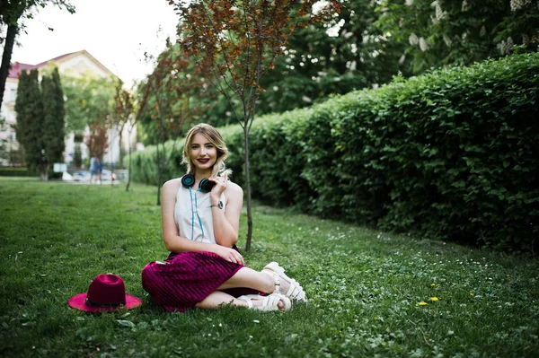时尚和美丽的金发模特女孩在时尚的红色天鹅绒天鹅绒裙子 白色上衣和帽子 坐在绿色的草地上 在公园里的电话和耳机 — 图库照片