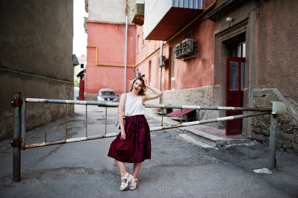 Fashionable and beautiful blonde model girl in stylish red velvet velour skirt, white blouse, hat and handbag, posed against barrier.