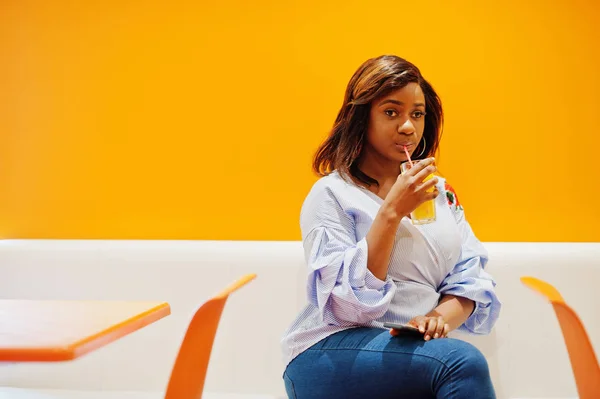 Mujer africana sentada en la cafetería contra la pared naranja beber pineappl — Foto de Stock