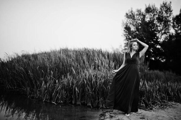 Blonde sensual barefoot woman in red marsala dress posing against lake with reeds.