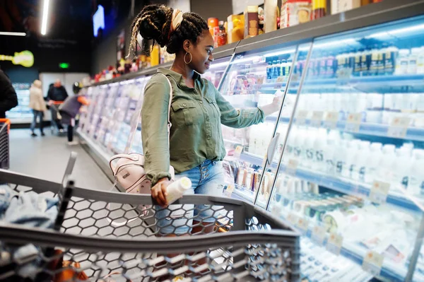 African woman with shopping cart choose yogurt bottle from fridge at supermarket.