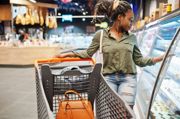 African woman with shopping cart choose yogurt bottle from fridg