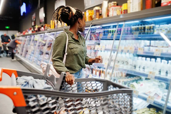 African woman with shopping cart choose yogurt bottle from fridg — Stock Photo, Image