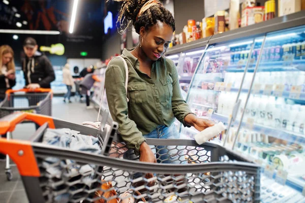 African woman with shopping cart choose yogurt bottle from fridg