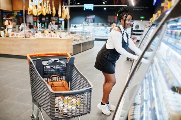 African woman with shopping cart choose yogurt bottle from fridg — Stock Photo, Image