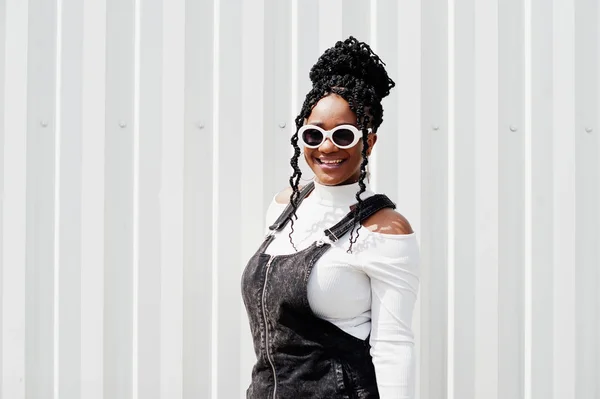 African woman in denim overalls skirt, white sunglasses posed ag