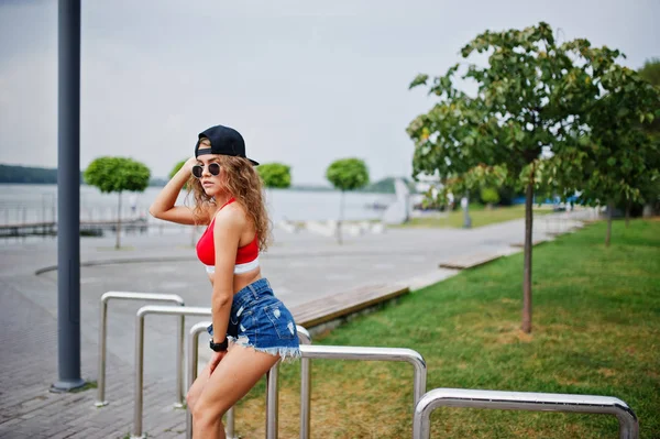 Sexy curly model girl in red top, jeans denim shorts, cup sungla
