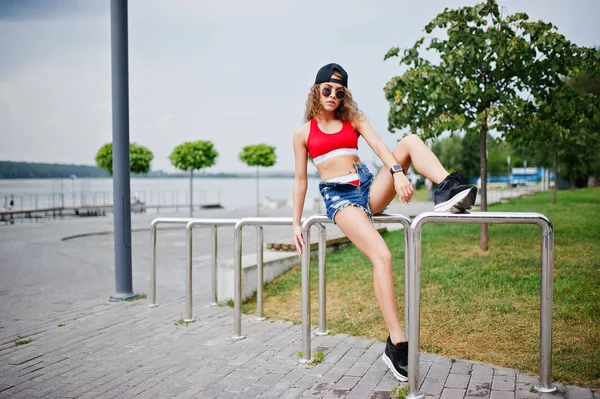Sexy chica modelo rizado en top rojo, jeans pantalones cortos de mezclilla, sungla taza —  Fotos de Stock