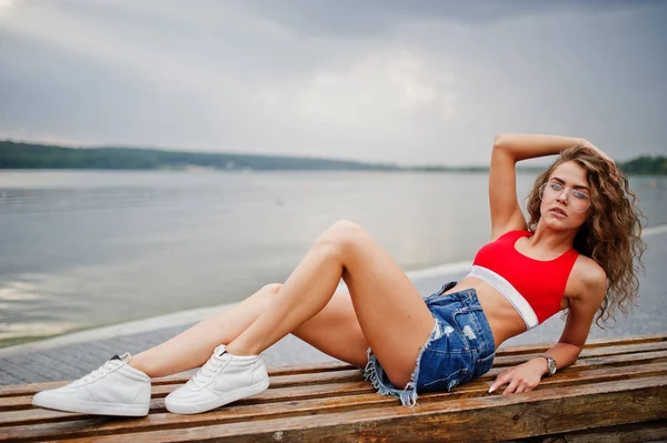 Sexy chica modelo rizado en top rojo, jeans pantalones cortos de mezclilla, gafas —  Fotos de Stock