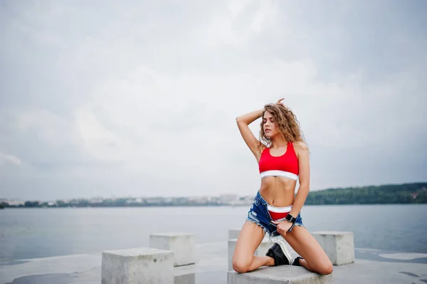 Sexy chica modelo rizado en top rojo, pantalones vaqueros vaqueros y zapatillas de deporte —  Fotos de Stock