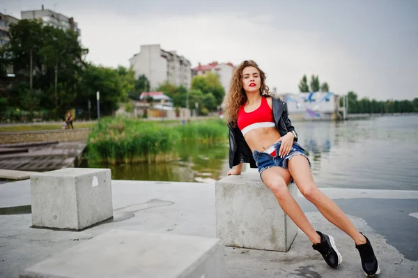 Сексуальная кудрявая девушка-модель в красном топе, джинсовых джинсовых шортах, кожаных джинсах — стоковое фото