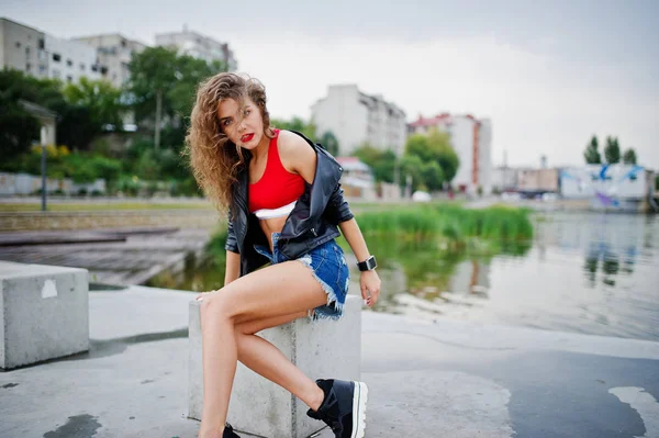 Sexy curly model girl in red top, jeans denim shorts, leather ja