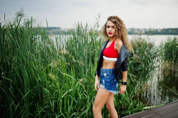 Sexy curly model girl in red top, jeans denim shorts, leather ja
