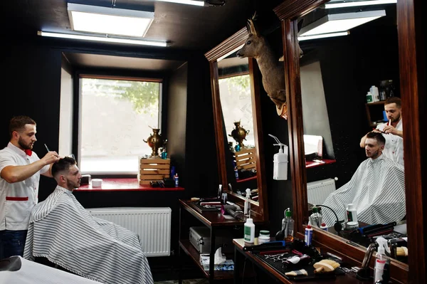 Young bearded man getting haircut by hairdresser while sitting i