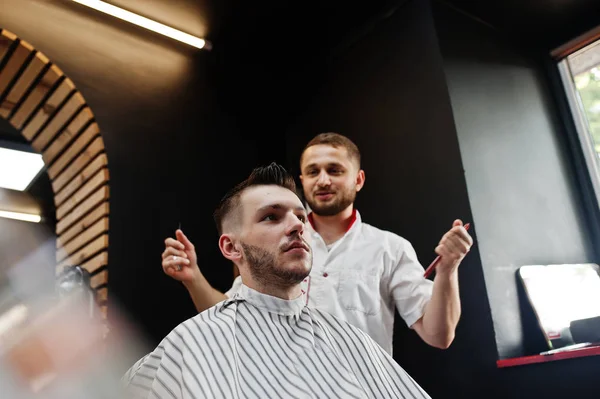 Young bearded man getting haircut by hairdresser while sitting i