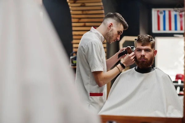 Young bearded man getting haircut by hairdresser while sitting i