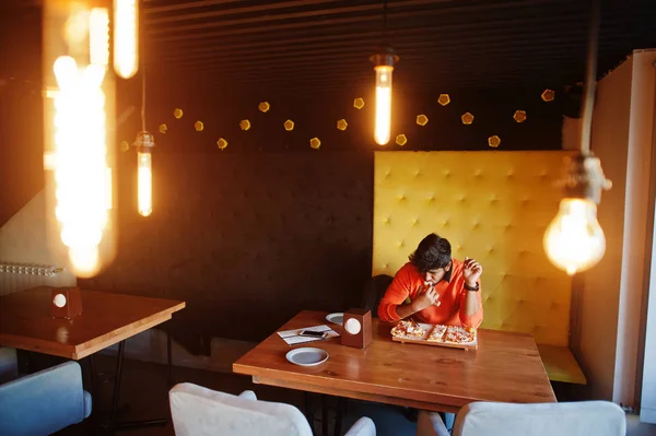 Уверенный молодой индиец в оранжевом свитере сидит в пиццерии — стоковое фото