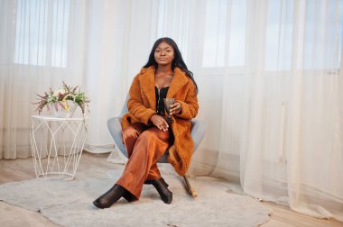 Turuncu montlu şık Afro-Amerikan kadın oturma odasında poz verdi.